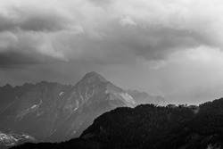 A grayscale shot of rocky mountains covered in trees under a cloudy sky in the countryside