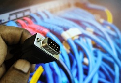 Male end of a VGA Cable having nine pins. A person holding the cable in front of a network switch