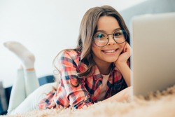 Mirthful teenager wearing glasses lying on the bed in front of a laptop with her hand touching the cheek