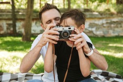 Photo of young father sitting with his little son outdoors in park nature holding camera photographing.