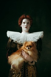 My best friend. Medieval redhead young woman in golden vintage clothing as a duchess holding puppy and standing on dark green background. Concept of comparison of eras, modernity and renaissance.