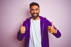 Young indian man wearing purple sweatshirt standing over isolated pink background success sign doing positive gesture with hand, thumbs up smiling and happy. Cheerful expression and winner gesture.