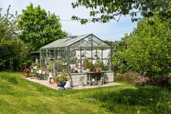 Beautiful greenhouse glass house in the garden yard near the villa. There are lots of pots with blooming blossom colorful flowers. Landscape garden design. Greenhouse for growing plant seedlings.