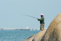 An unknown man standing fishing on a rock in the middle of the sea, industrial estate background.