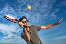 Man Holding A Paddle Tennis Racket Hitting The Ball On A Blue Background. Young Sportsman Playing Tennis On The Beach.