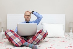 Man working with a laptop on a bed dressed in a shirt and tie and pajama pants and with his hand over his head with a worried expression. Teleworking concept