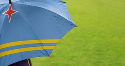 Aruba flag umbrella. Close up of printed umbrella over green grass lawn background. Rainy weather forecast. Climate change and global warming concept.