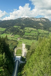 A view from the top of the ski flying hill in Bad Mitterndorf, Austria captured in summer. The ski jump is surrounded by thick forest from each side. High mountains in the back. Sunny and warm day.