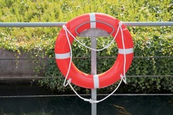 A single isolated orange red help lifebuoy handing with a thick light beige cloth rope on a metal pipe next to a river during summer, emergency life saving guard item