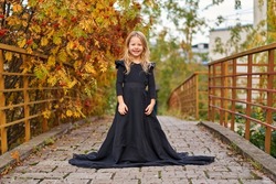 Happy laughing little girl on autumn trail in long magic black fashion dress. Halloween in gorgeous princess dress. Retro smile. Place for text. Laughing funny child girl in witch costume. road bridge