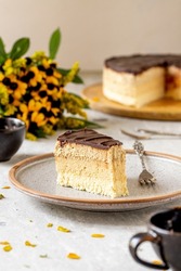 The layered creamy no-bake cake called in Serbia, Pesak torta, with ground biscuit, coconut, and chocolate layers served on a plate with yellow flowers in the back on the table
