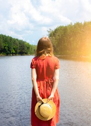 Beautiful girl with a straw hat near the lake. Amazing summer nature. Travel concept. Inspiration for wanderlust. Woman in linen red dress. Eco-friendly materials for clothes.