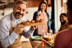 Happy mid adult man serving healthy bruschetta while having a meal with friends at home. 