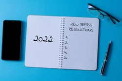 Selective focus of pen, glasses, smartphone and notebook written with 2022 new year's resolutions on blue background.