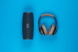 A pair of headphones next to a black speaker on blue background, shot from above.