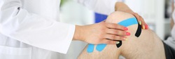 Traumatologist glues blue and black tape on patient's sore knee in clinic closeup. Trauma recovery kinesiotherapy concept.
