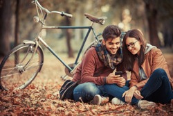 Young couple sitting outdoors at the park on beautiful autumn day.They sitting leaves and using smart phone.