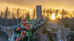 Wooden grave cross with artificial flowers at sunset. Orthodox traditional cemetery in snowy winter. Space for text.