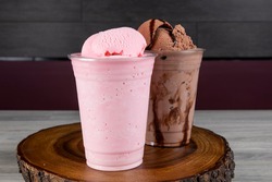 A takeout glass of cherry or strawberry milkshake with a glass of chocolate milkshake with large scoops of ice cream on a wooden tray