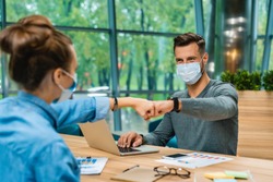 Two young caucasian colleagues joining fists in medical masks against Covid19 working in office