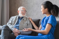 Caring african-american nurse talks to old elderly senior patient holds his hand sit in living room at homecare visit provide psychological support listen complains showing empathy encouraging.