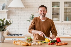 Handsome mature middle-aged man husband father cooking vegetable salad in the kitchen at home, preparing vegetarian food meal cutting bell pepper on cutting board. Homemade meal