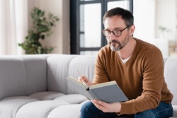 Relaxed pensive middle-aged mature man father husband author freelancer tutor adult student lecturer reading book at home, preparing for exams at university, enjoying novels at leisure time.