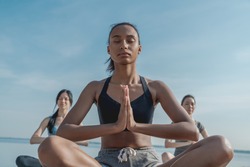 Low angle view of young woman on yoga class doing meditation lotus pose on the beach. Yoga training class with coach meditating outdoors together. Serene people.