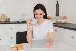 Happy young caucasian woman student freelancer business woman using digital tablet while having breakfast in the morning, reading news, surfing social media, e-learning, watching videos and movies