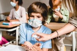 Young teacher using sanitizer with her pupils schoolchildren kids students at lesson class in school wearing protective medicine masks against Covid19 coronavirus. School during pandemic