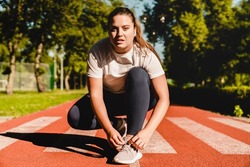 Plump plus-size body positive woman athlete tying sporty shoes, active wear, sneakers for training workout jogging running in stadium outdoors.