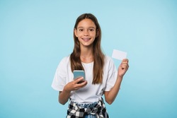 Smiling cheerful caucasian teenage girl student pupil schoolchild holding credit card and smart phone for shopping online, Internet banking, bank accounts isolated in blue background