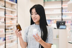 Female asian customer client looking for medications, medicines, comparing two jars with drugs, pills, vitamins, antibiotics, painkillers, choosing between two products in pharmacy drugstore