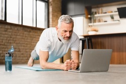 Mature middle-aged athlete man standing in plank position in sporty clothes on fitness mat, watching video training tutorial online, vlogging and blogging at home on lockdown