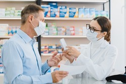 Male customer visitor giving medical prescription to female pharmacist in medical mask against coronavirus, while druggist advising new remedy pills antibiotics
