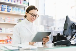 Chemist druggist pharmacist using digital tablet for checking looking for active substance, side effects of drugs remedy pills medicines online standing at cash point desk in pharmacy drugstore