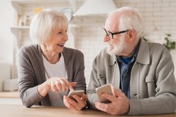 Smiling caucasian senior elderly couple grandparents spouses using smart phones cellphones together, surfing social media, e-banking, e-commerce at home kitchen, online shopping on application