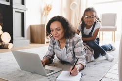 Busy african-american mother freelancer working on distance remotely on laptop, trying to concentrate while her naughty preteen daughter disturbing her. Maternity leave concept. Stay at home