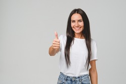 Positive caucasian young girl woman in white T-shirt showing thumb up looking at camera, checking for good quality recommendation isolated in grey background. Well done! Great job!