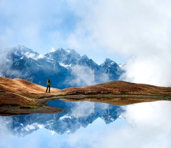 Lake with reflection of the rocky ridge covered with snow and young woman enjoying the view. Beautiful landscape from Svaneti, Georgia, Europe.
