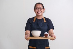 A waitress wearing a black apron over a black T-shirt serves a large cup of coffee and a chocolate muffin sprinkled with powdered sugar, on a wooden tray. Happy and friendly expression.