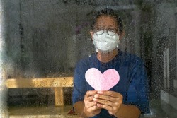 Close-up: A heart-shaped pink paper is held by a woman wearing mask who is isolated at home due to covid-19 to say I love you.