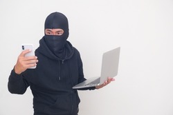 A male thief in a black mask and hood is in action; carries a laptop and hold a cellphone.