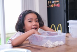 A little girl enjoys popcorn served on a wooden dining table; smiling, glancing, happy expression. 