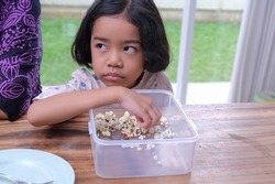 A little girl enjoys popcorn served on a wooden dining table; frowning, glancing, unhappy, upset. 