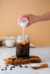 Ice coffee in jar with cream poured over and coffee beans on table. Cold summer drink.