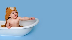 Happy baby toddler boy with hood bathrobe shows his hand in a white tub on a studio blue background. A smiling child at the age of one year, copy space