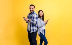 Portrait of smiling couple dressed in casuals using smart phones against yellow background. Cheerful young man and woman standing back to back and messaging online on social media