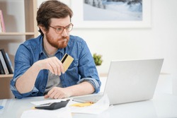 Troubled bearded guy holding credit card and using laptop
