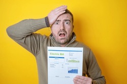 Shocked man reading some bills energy expenses isolated on yellow background